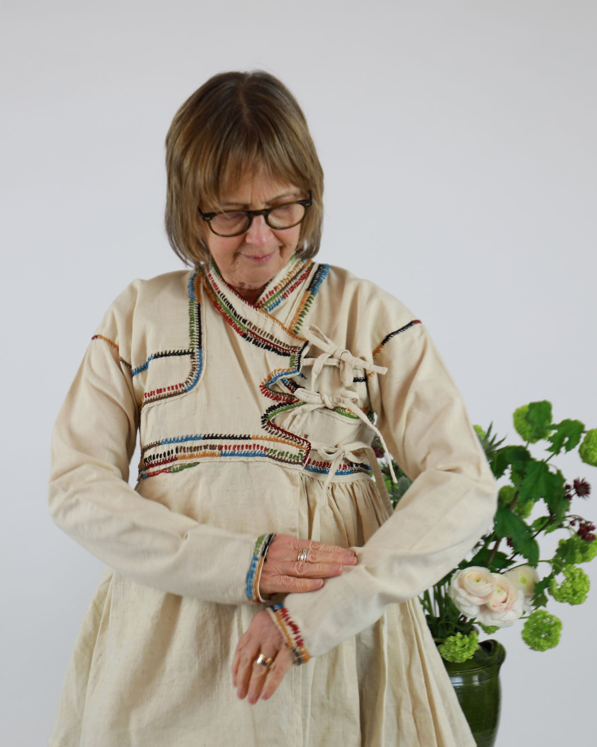Woman wearing a traditional outfit with a colorful scarf, standing next to a vase of flowers on a white background